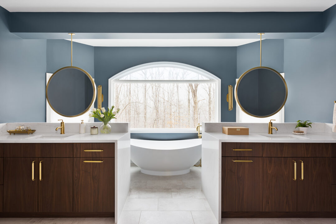 Clifton VA primary bathroom remodel featuring walnut double vanities flanking a freestanding soaking tub beneath a large picture window overlooking a wooded backyard.